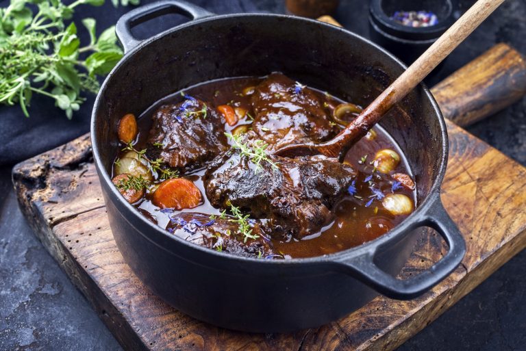 Traditional German braised beef cheeks in brown red wine sauce with carrots and onions offered as closeup in a cast iron Dutch oven on an old rustic cutting board
