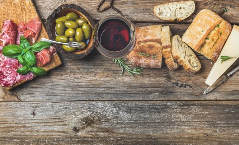 Wine snack set. Glass of red wine, green mediterranean olives, prosciutto, salami and aged cheese over rustic wooden background. Top view, copy space